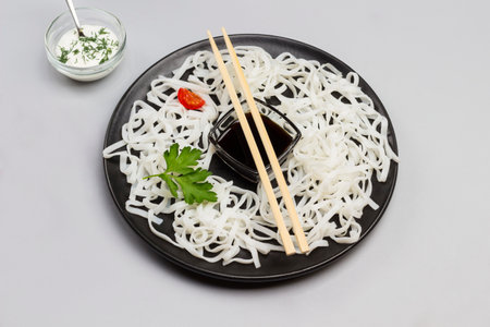 Rice noodles with sauce, bamboo sticks on black plate. Cherry tomatoes in bowl with white sauce on table. Gray background. Top view.の写真素材