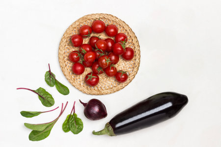 Cherry tomatoes in wicker plate. Eggplant, broccoli and chard leaves on table. White background. Flat lay.の写真素材