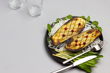 Baked eggplant with greens and rice noodles on black plate. Fork and knife. Sauce in glass bowl. Top viewの写真素材