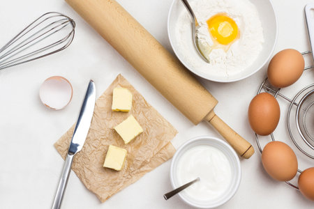 Brown eggs on metal stand. Butter and knife on paper. Egg yolk with flour and spoon in bowl. Milk in bowl and rolling pin on table. White background. Flat layの写真素材