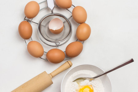 Brown eggs on metal stand. Egg yolk with flour and spoon in bowl. Milk in bowl and rolling pin on table. White background. Flat layの写真素材