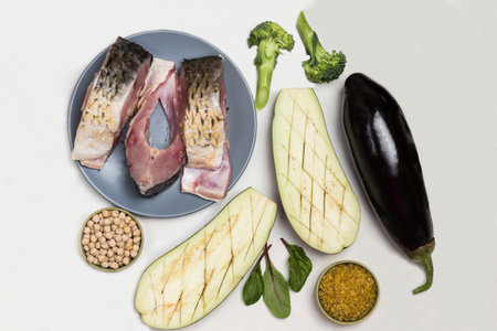 Pieces of river carp fish in plate. Eggplant, greens and groats on table. White background. Flat layの写真素材