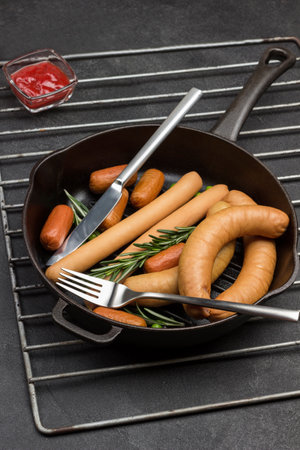 Sausages and sprigs of rosemary with knife and fork in frying pan on metal grate. Tomato paste, mustard and horseradish on table. Black background. Top viewの写真素材