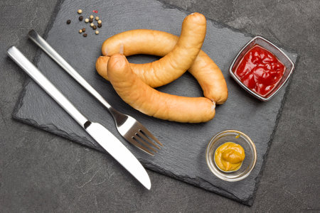 Sausages on slate board. Knife and fork. Mustard and tomato paste in glass bowls. Black background. Flat layの写真素材
