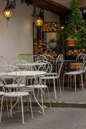White tables and chairs on the summer terrace of the cafe. Lanterns are burning above the tables. Chopped firewood is on the shelves.の写真素材