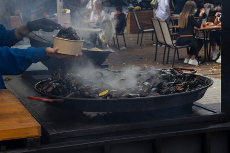 Grilled mussels on a large frying pan. Light steam. Hands scooping mussels into a container. Street food.の写真素材