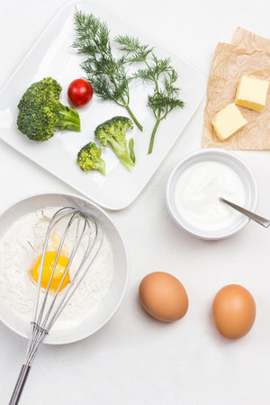 Broken egg with flour in bowl. Whisk on bowl. Butter on paper. Broccoli, tomatoes and dill in plate. White background. Flat layの写真素材
