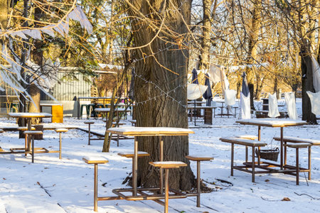 Tables and benches covered with snow on the winter terrace of a cafe. Bare trees are decorated with white flags. Cloth napkins hang on a rope between the trees.の写真素材
