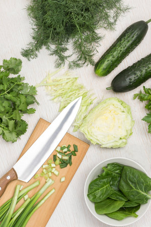 Spinach leaves in white bowl. Green onions and knife on cutting board. Cabbage, cucumbers and greens on table. White background. Top viewの写真素材