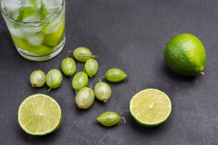 Slices of lime and gooseberries on table. Two glasses of cold drink with lime, mint and ice. Black background. Top viewの写真素材
