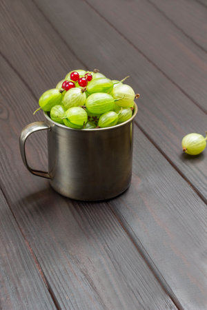 Metal mug with green gooseberries and red currants on top. Dark wooden background. Top viewの写真素材