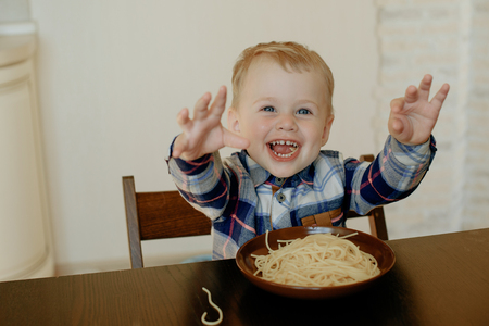 A cheerful boy of two years eating spaghetti with his hands at home in the kitchenの写真素材