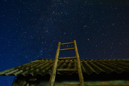 Village. Old wooden ladder leaning against the barn with a slate roof in the night star sky. The staircase leads to the sky.の写真素材