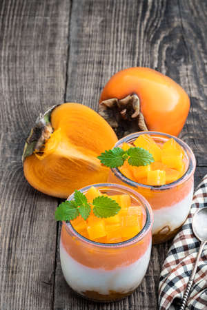 White fruity yogurt with persimmon slices in glass jars on wooden tableの写真素材