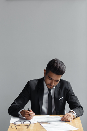 Handsome young business man writing, businessman working with documents sign up contract, sitting at the desk at office, wear elegant suit and tie isolated over grey backgroundの写真素材