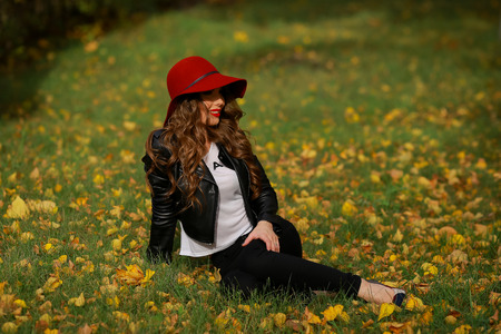 Beautiful woman in red hat sitting on the grass in the park in autumnの写真素材