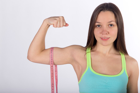Young strong woman in sportswear measures biceps muscle with centimeter tape, white background, front viewの写真素材
