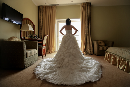 bride in a white dress with a long train standing at the window in hotel.の写真素材