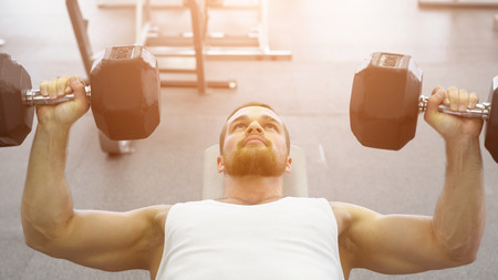 bodybuilder does bench press using dumbbells at the gym, sunlight. Muscular man exercising in the gym, close upの写真素材