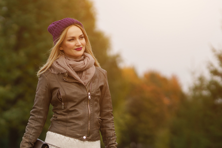 Stylish autumn. Fashionable pretty blond woman in cap, leather jacket and with handbag posing in autumn park, sunlightの写真素材
