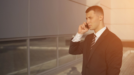 Attractive manager in formal suit talking on phone while standing on background of high-rise building on sunny day. sunlight, copyspaceの写真素材