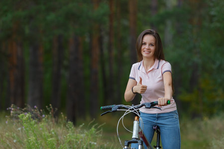 Happy girl shows thumb up, riding on bicycle in pine forest, sport leisure. Cyclist walk in park,...