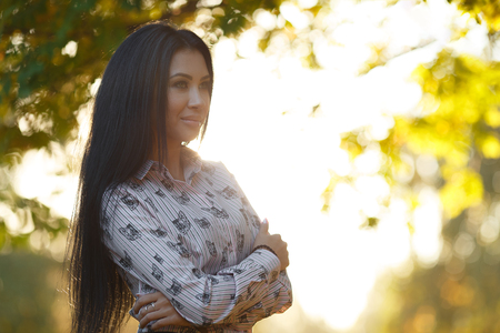 Portrait of beautiful stylish young brunette with long hair standing in sunny summer park against background of green blurry treesの写真素材
