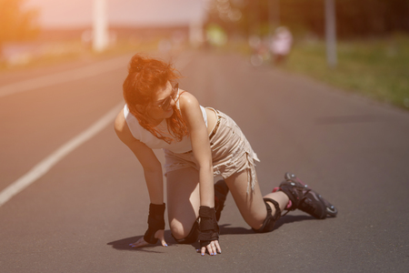 Charming red-haired girl in sunglasses and protective gear rides on roller skates and fell on the road on sunny day, sunlightの写真素材