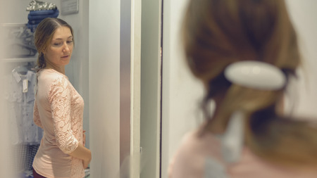 Pretty girl looks at her reflection in the mirror while trying on clothes in fashion store while shoppingの写真素材