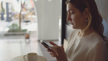 Side view - beautiful young woman typing text on smartphone sitting at table in cafe on the...