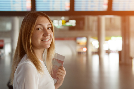 beautiful young girl stands and looking at camera on the background of scoreboard with the notice. woman with passport and ticket in hands, sunlightの写真素材
