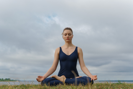 Young woman in sportswear sitting in yoga pose, cloudy sky on background. Yogi training, outdoor meditation, healthy lifestyleの写真素材