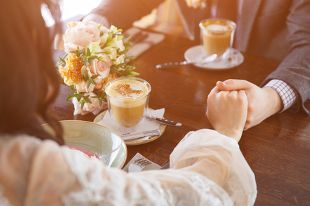 Bride and groom in a luxurious restaurant holding hands and drinking a cup of coffee latte, sunlightの写真素材