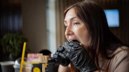 side view of young girl in black gloves eating a burger in cafe.