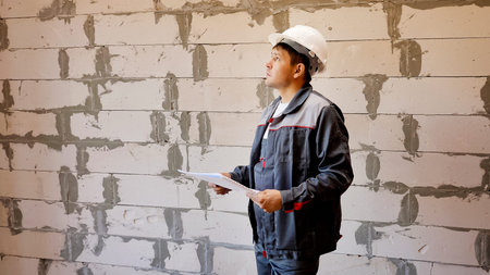 Adult man in hardhat reading papers and looking around in unfinished room with bare brick wall on...