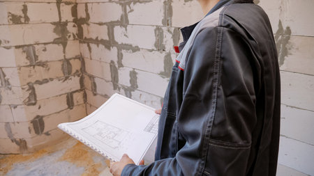unknown man in uniform working on site of new house standing in empty room with paper plans.