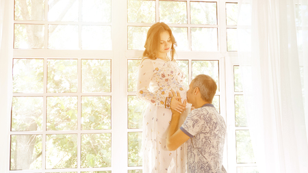 Side view of happy man kissing belly of crop pregnant woman in dress standing in lightの写真素材