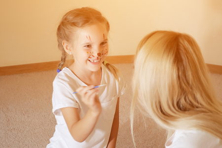 Excited little girl with painted snout having fun with mother drawing on her face with paintbrushの写真素材