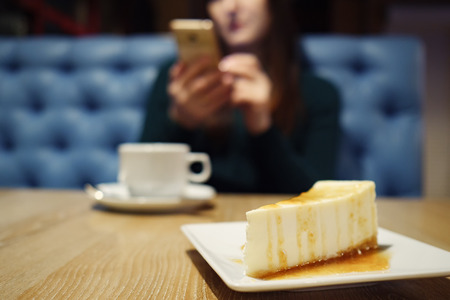 blurred of unknown girl with phone and cup of coffee. Selective focus on the cheesecake, close upの写真素材