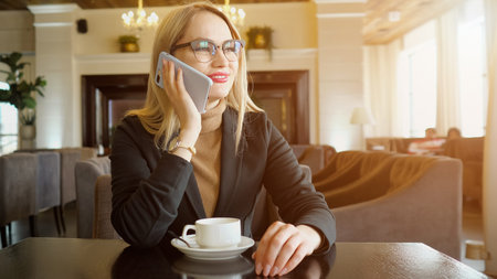 Smiling young woman having friendly phone conversation while drinking coffee in cafe, sunlight