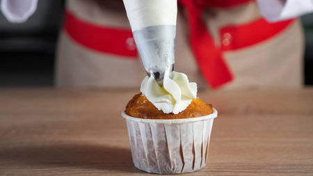 Pastry chef decorates cupcake in paper cups white cream with pastry bag. Hands close-up. Baking and...
