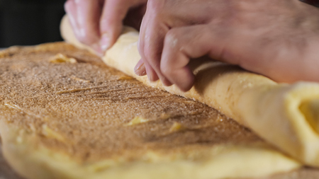 Pastry chef rolls the dough with cinnamon, sugar and butter into a roll on the table, selective focus. close-up.の写真素材