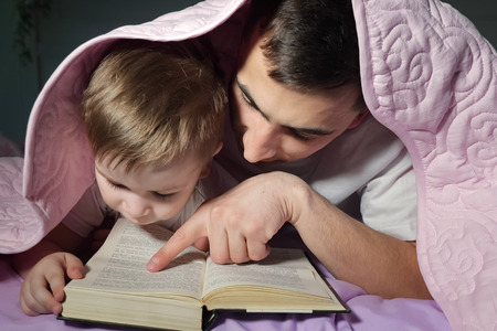 Portrait of dad teaches his little son to read a book hiding under the blanket in bed before bedtime.の写真素材