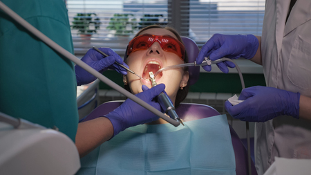 Dentist is taking care about patients tooth with black cavity on it using electric dental brush. Nurse with saliva ejector near the patient is helping to doctor. Womans portrait in dental chair.の写真素材