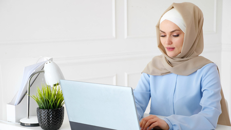 Portrait of young muslim woman in hijab and traditional dress is typing on laptop working in office. Arabian woman at work on her workplace.の写真素材