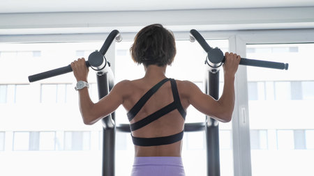 Slim sporty woman is doing pull-ups on horizontal bar with wide grip in gym. She is in sportswear in...