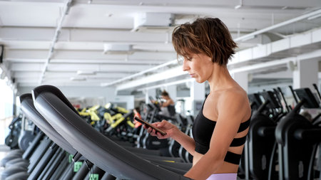 Mature sporty woman is training on treadmill in gym and looking in mobile phone. She is walking in...