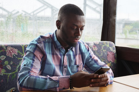 Bearded black young man looking at mobile phone. Afro american man is browsing smartphone sitting in...