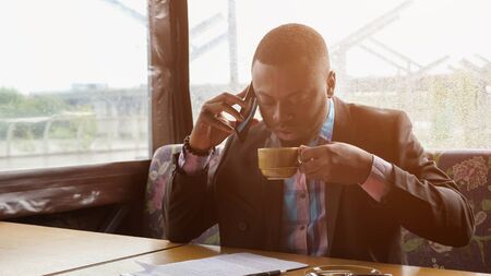 Black guy is talking on smartphone. Afro american businessman is calling on mobile phone sitting in summer tent cafe. He is drinking a cup of coffee at table, sunlight. He wears shirt and suit jacket.の写真素材