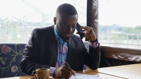 Black guy is talking on smartphone. Afro american businessman is calling on mobile phone sitting in summer tent cafe. He is drinking a cup of coffee at table. He wears shirt and suit jacket.の写真素材